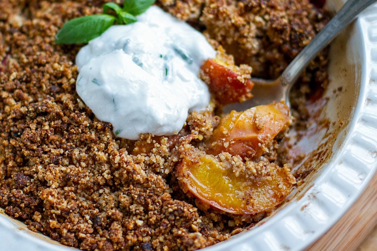 Peach crisp in a baking dish with a spoon and a dollop of basil coconut yogurt on top