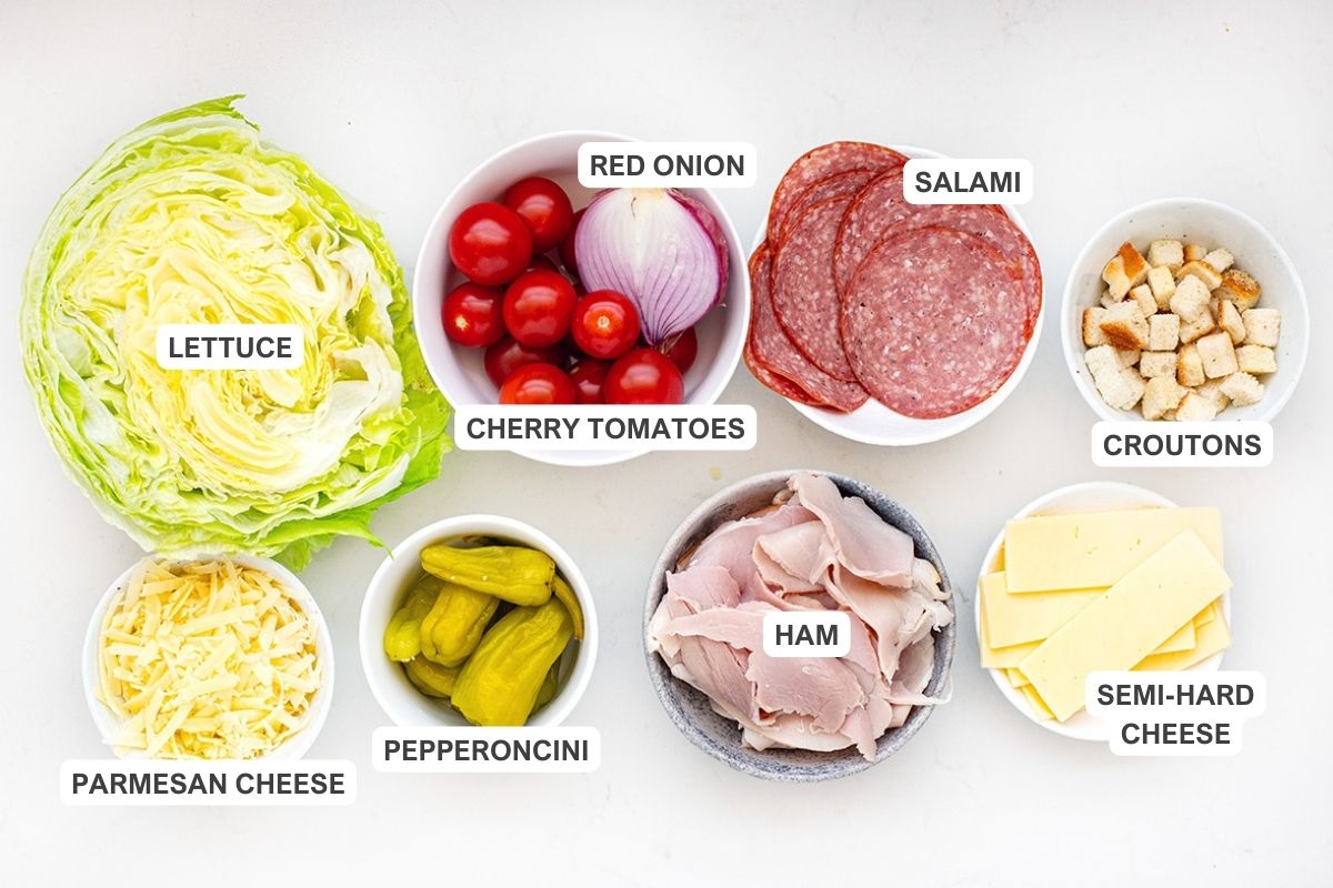 Ingredients for the recipe including lettuce, cherry tomatoes, red onion, salami, croutons, parmesan cheese, pepperoncini, ham, and semi-har cheese.