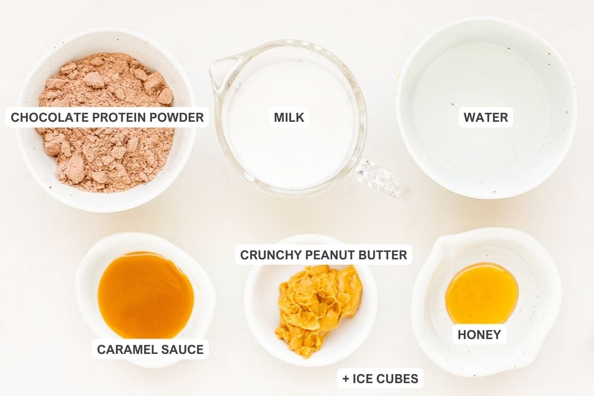 Ingredients for the recipe including chocolate protein powder, milk, water, caramel sauce, crunchy peanut butter, honey, and ice cubes.