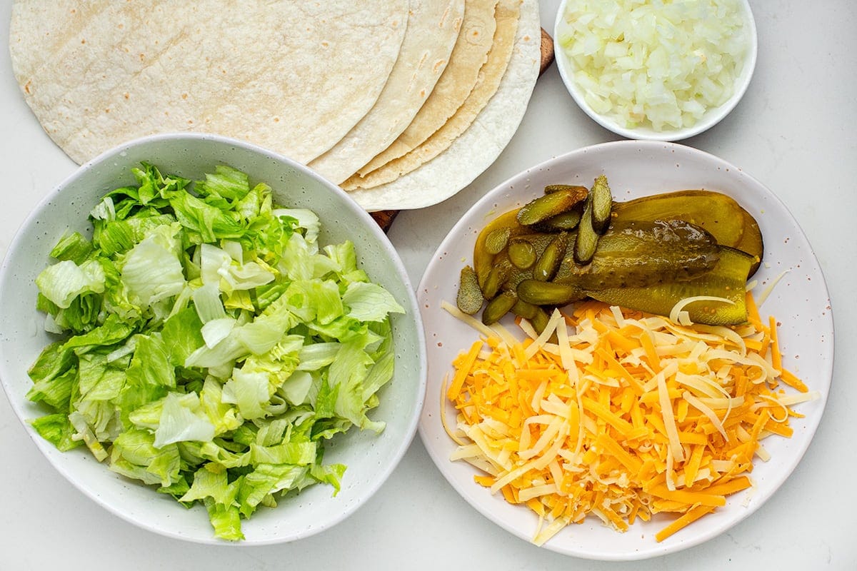 Ingredients for the recipe including high-protein wraps, shredded iceberg lettuce, shredded cheddar cheese, sliced pickles, and chopped onion.